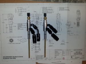 Two completed pickup tube assemblies for a Paxton SN60 oil cooling system placed on top of detailed SolidWorks engineering schematics. The tubes feature precision-machined aluminum heads, brass dipstick-style rods, and black AN fittings for fluid connections. The background showcases labeled dimensions and design notes, emphasizing technical craftsmanship and design accuracy.
