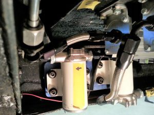 Close-up of the installed foundation and oil circulation pumps for the Paxton SN60 supercharger cooling system. The setup includes braided stainless-steel lines with AN fittings, mounted to a precision-machined aluminum foundation. The pumps and connectors are securely fastened, demonstrating a robust and professional installation within the vehicle's underbody framework.